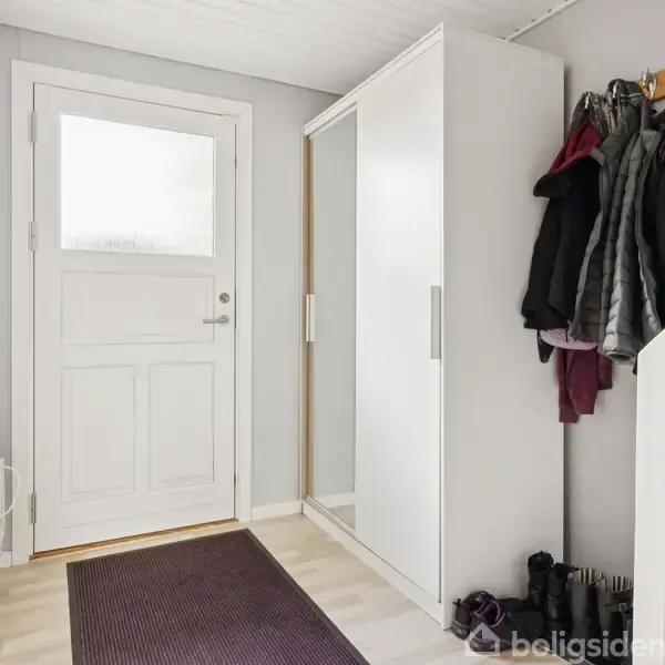 Yderdør står ved siden af et stort, hvidt skab. Jakker hænger på knager ved siden af døren. Et par sko står på gulvet, og en mørk løber er placeret på det lyse parketgulv.