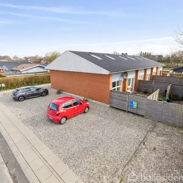 Rødt murstenshus med fladt tag står på en grusparkeringsplads. En rød og en grå bil er parkeret foran huset. Omgivelserne viser en vej og andre bopæle.