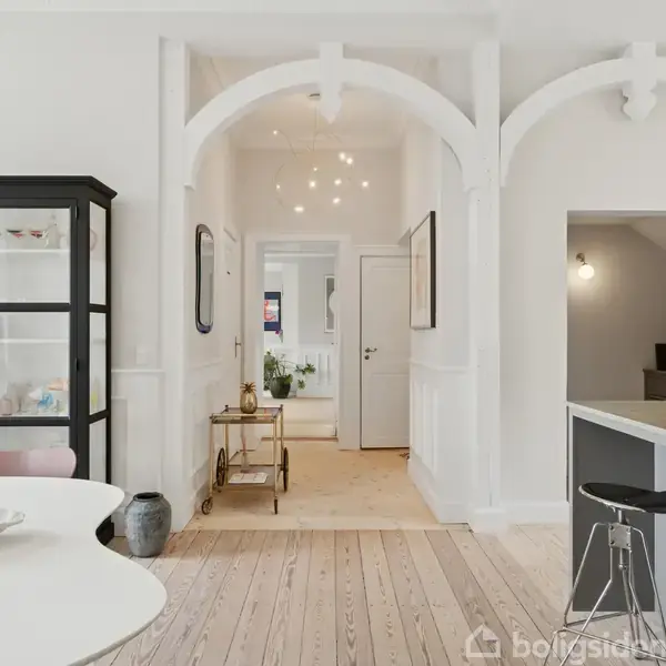 Sort vitrineskab står i en lys spisestue med trægulv. En barvogn står i midten under buede åbninger, mens barstole står ved en grå køkkenø.