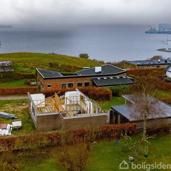 Bygning under konstruktion står i en frodig have med udsigt over en stille fjord. Omkringliggende huse og havn er delvist skjult af tåge i baggrunden.