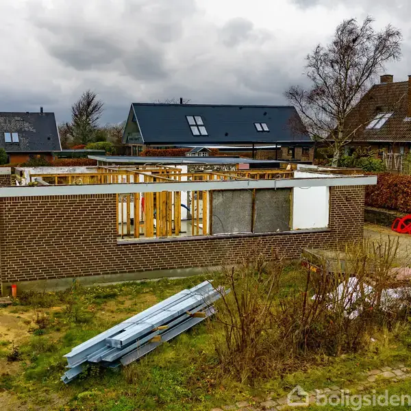Bygning under konstruktion med brune murstensvægge og delvist åbent interiør. Omkringliggende have er ujævn med græs og buske. Bagved ses flere huse med gavle og skyer på himlen.