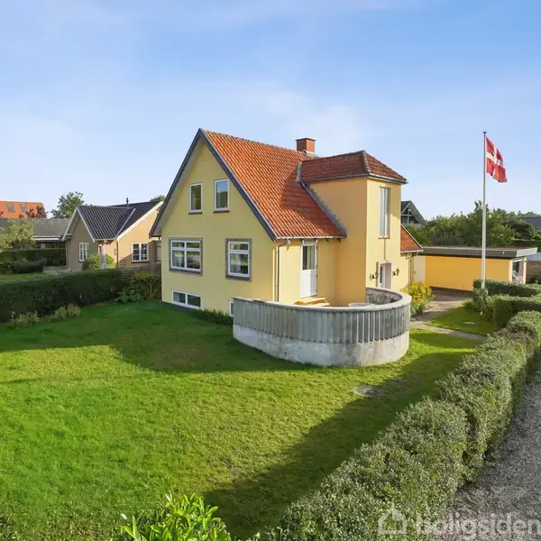 Et gult hus med rød tag står i en velholdt have. Der er en flagstang med Dannebrog, og en indkørsel af grus. En bænk står i græsset nær hække.