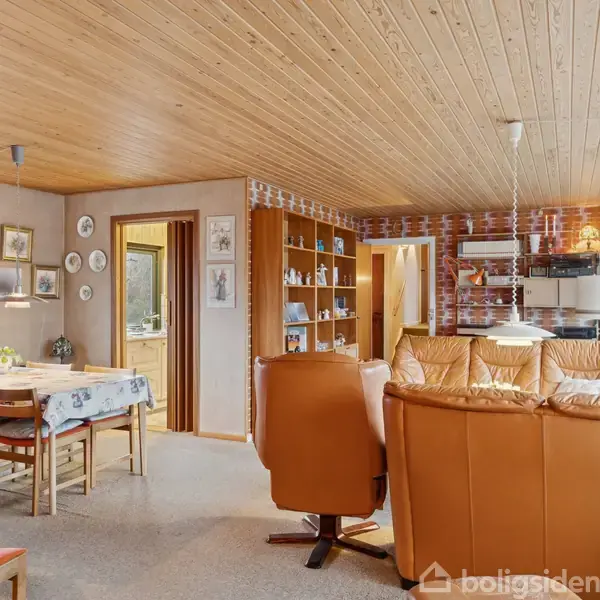 Stue med rustikke lædersofaer, der vender mod en åben reol og spisebord. Vægge af røde mursten og træloft skaber en varm atmosfære. Vinduer giver udsigt til haven.