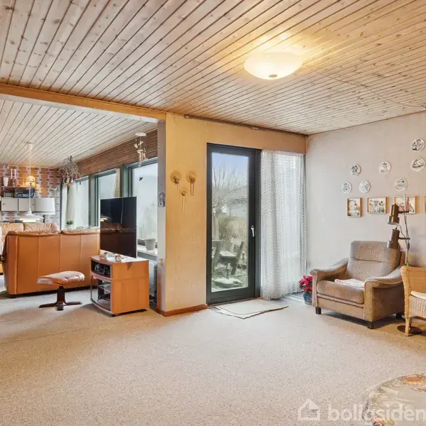 Stue med tæppebelagt gulv og træloft. To stole til højre ved siden af en glasdør. Billeder og dekorationer pryder væggen. Køkken ses i baggrunden.