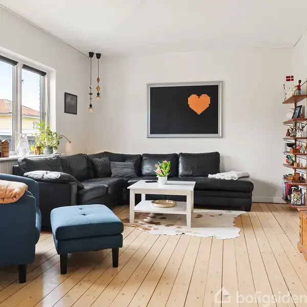 En stue med en mørk hjørnesofa omkring et hvidt sofabord. Et blå lænestol og skammel står foran store vinduer med planter. En orange hjerteplakat hænger på væggen.