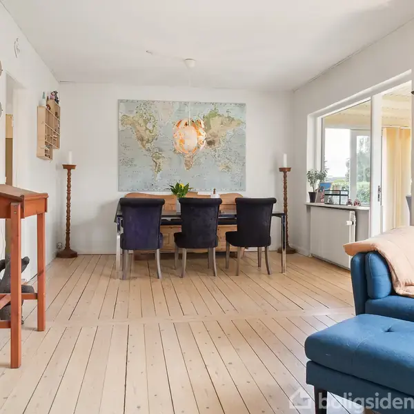 Blå sofa står i stue med lyse trægulve, et spisebord med sorte stole og en verdenskortplakat på væggen. Rummet har store vinduer og forskelligt interiør som møbler og dekorationer.