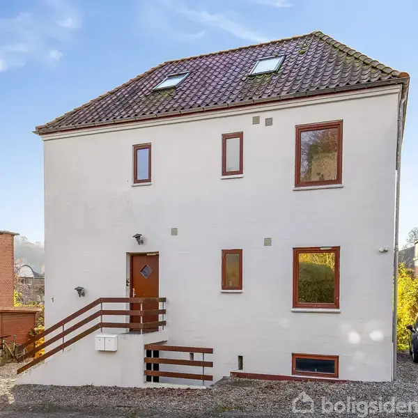 Hvidt hus med skråt tag og brune vinduesrammer. Ingen aktivitet ses. Omgivet af grusvej, bil og grønne buske i et beboelsesområde.