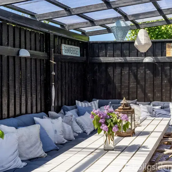Langt træbord med blomster i vase og lanterner, omgivet af polstrede bænke med puder, står i en sortmalet, overdækket terrasse med gennemsigtigt tag og hængende lamper. Skilt: "Svalehuset".