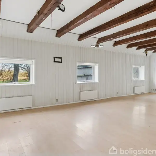 Et lyst rum med synlige træbjælker i loftet, tre vinduer langs væggen og lyst trægulv. Udenfor ses træer, og indenfor er rummet sparsomt møbleret.