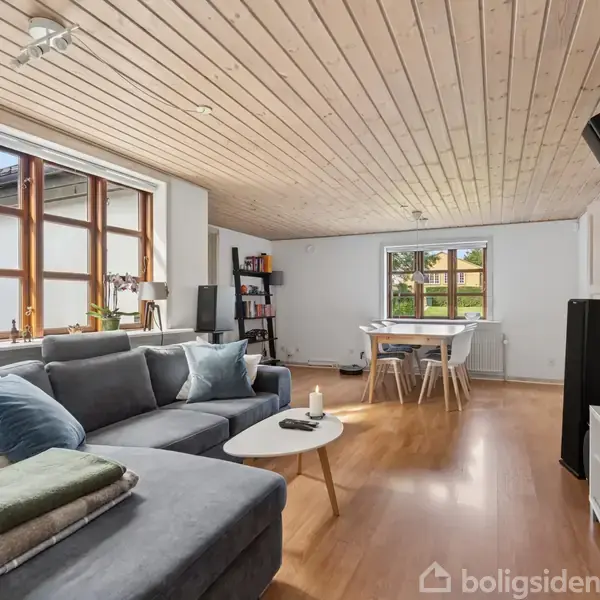 Grå sofa står i en stue med trægulve, flankeret af et sofabord og reol. Et spisebord med stole står ved vinduet. Loftet er træbeklædt.