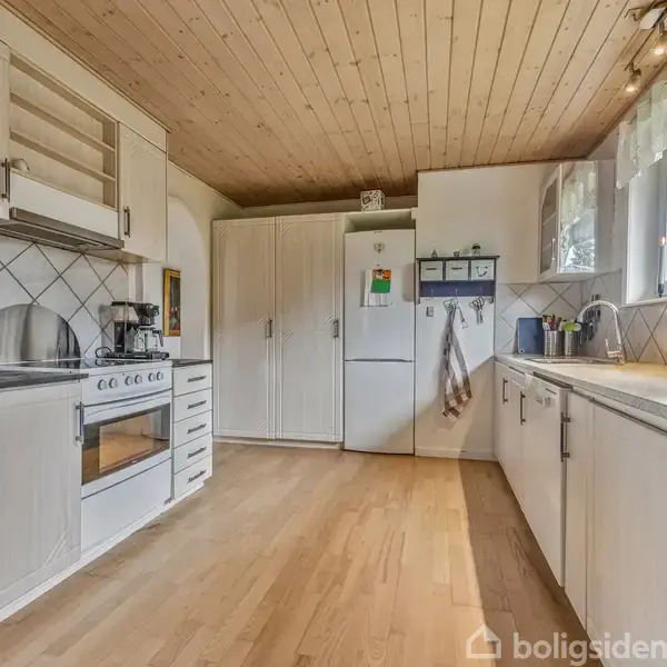 Et lyst, veludstyret køkken med hvide skabe og apparater. En stor bordplade strækker sig langs væggen under et vindue med udsigt til det fri. Træloft giver en varm atmosfære.