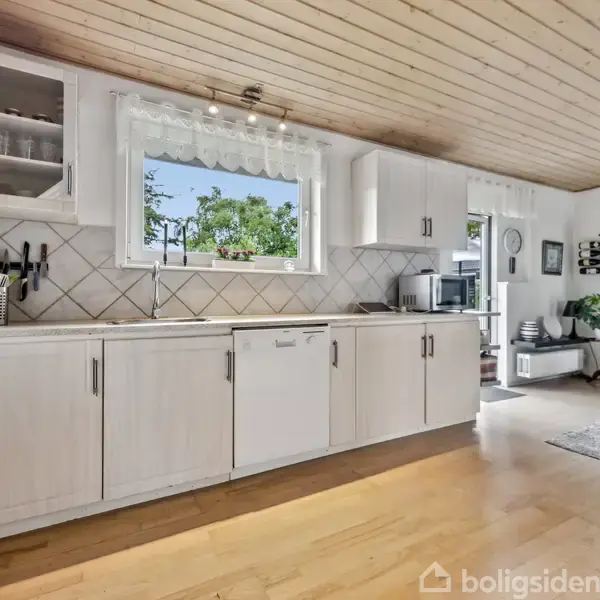 Køkken med hvide skabe og opvaskemaskine, beliggende i en lys stue med trægulv. Et vindue over køkkenbordet viser udsigt til grønne træer udenfor. Rundt spisebord ses til højre.