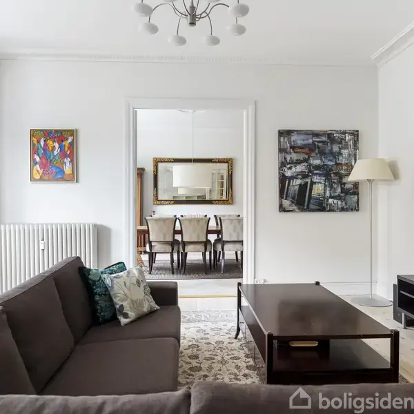 En stue med en brun sofa foran et sofabord, en væghængt fladskærm, kunst på væggene og en åben døråbning til en spisestue med spisebord og stole.