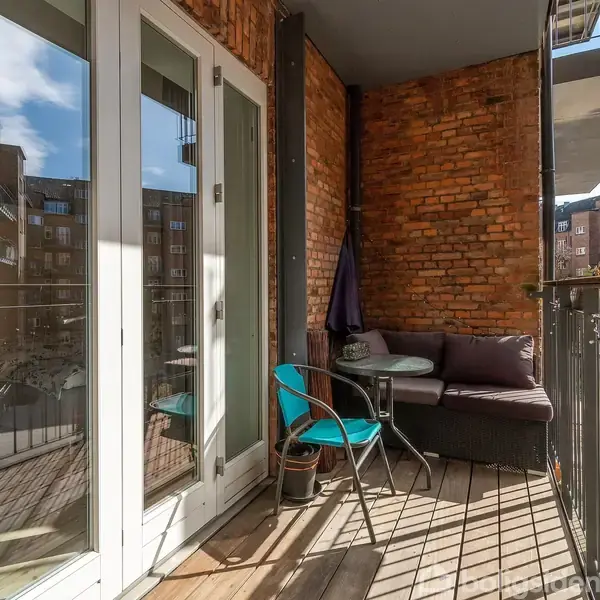En balkon med sofa og glasbord omgivet af murstensvægge. En blå stol står ved bordet. Store glasdøre fører til lejligheden. Bygninger kan ses i baggrunden.