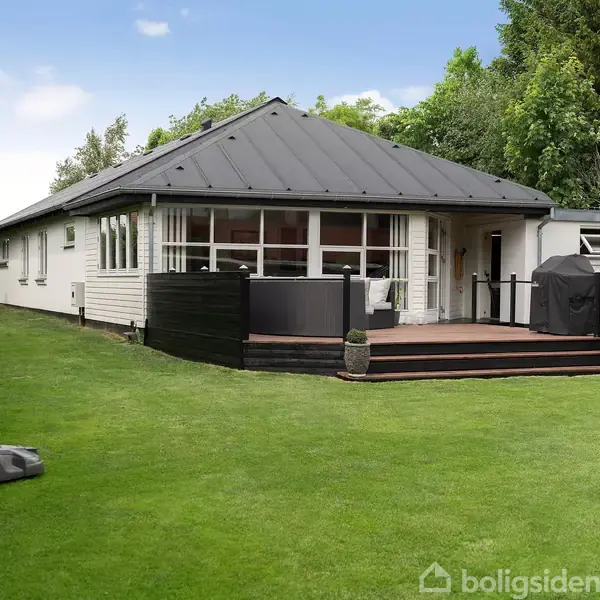 En hvid enkeltplansvilla med sort tag ligger i en frodig have med velplejet græs. En robotplæneklipper bevæger sig over græsplænen. Terrasse med sorte hegn og grill i baggrunden.