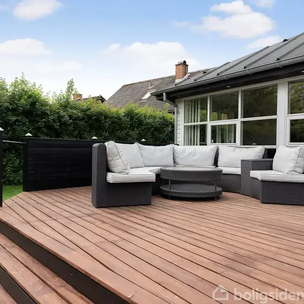 Udendørs terrasse med sorte møbler og grå hynder står på en træplatform. En rund ildsted er i midten. Omgivet af en grøn have og buske nær et vinduesfacadehus.