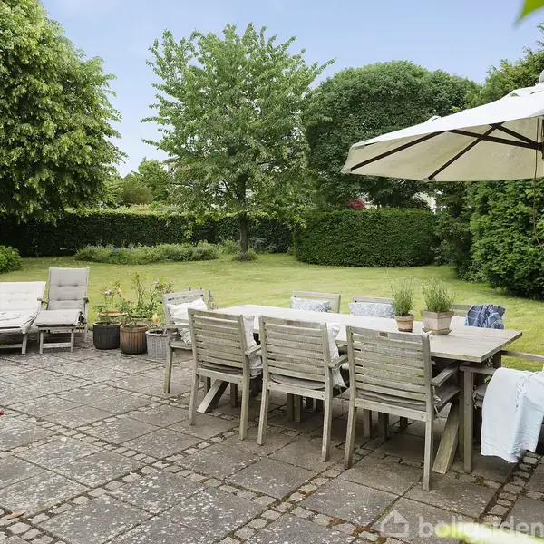 En træterrassemøbelgruppe står på en flisebelagt terrasse med en stor parasol over. Flere stole og liggestole omkranser bordet. Græsplæne og buske omgiver området i baggrunden.