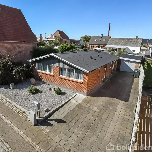 En murstensbungalow med mørkt tag står på en flisebelagt indkørsel. Haven er dekoreret med grus. Til venstre er en bil parkeret på vejen.