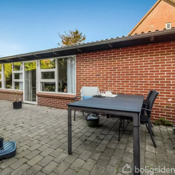 En sort havebord med stole står på en flisebelagt terrasse foran et hus med røde mursten og store vinduer. Planter omgiver terrassen, og himlen er blå.