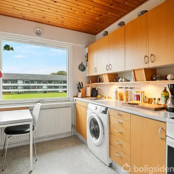 Vaskemaskine i køkken står under træskabsarrangement. Komfur og kaffemaskine ved siden af. Bord med to stole ved vindue med udsigt til grønne omgivelser og lejlighedskompleks.