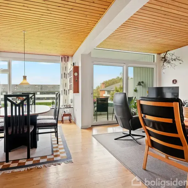 Spisebord med stole står på et tæppe, mens stole og sofabord er på gulvtæppe i en lys stue med trægulv og træloft. Store vinduer åbner mod en terrasse.