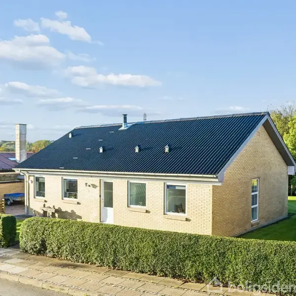 Et enkelt enfamiliehus med mørkt tag og lysegule mursten står stille i et forstadskvarter. Huset er omgivet af en klippet hæk og en velholdt græsplæne under en klar himmel.