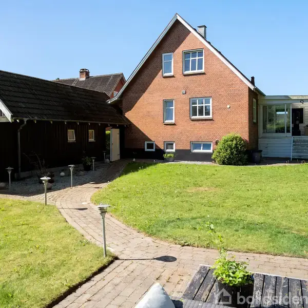 Rødt murstenshus med tilhørende carport i træ, stående i en grøn have med flisebelagt gangsti. I forgrunden ses et udendørs siddeområde med bord og planter.