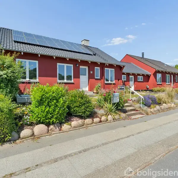 Et rødt murstenshus med flere vinduer står på en velplejet græsplæne. En terrasse ses foran huset. Det er under en blå himmel med enkelte skyer.