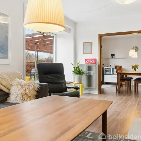 En stue med grå sofa og sort lænestol ved et træbord, oplyst af en gul lampe. I baggrunden ses en spisestue med et spisebord og stole.