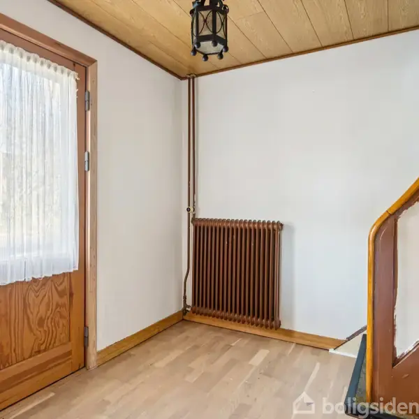 Dør med hvidt gardin står i et hjørne af et rum med trægulv og træloft. Til højre er en buet trappe med trægelænder. En radiator ses under vinduet.