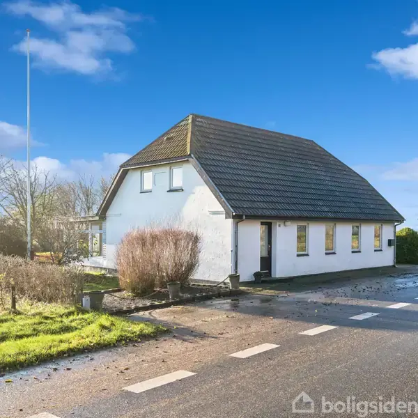 Et hvidmalet hus med mørkt tag står ved en vej. Solskin oplyser omgivelserne, og grønne græsarealer samt buske omringer huset. Flot blå himmel med få skyer ses i baggrunden.