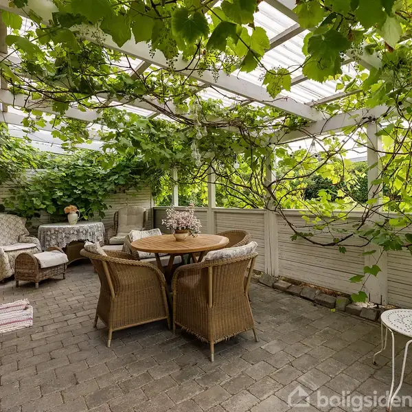 Rundt havebord omgivet af fletstole under en pergola dækket af grønne vinranker. I baggrunden ses en sofa og stole med hvidt mønster, og der står en lille hvid metalbord.