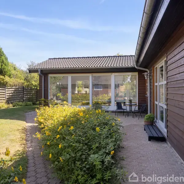 Hus med træbeklædning har en terrasse med bord og stole ved glasdøre. Gangstien er omkranset af gule blomster. Hegn og buske omkranser den græsbeklædte have.