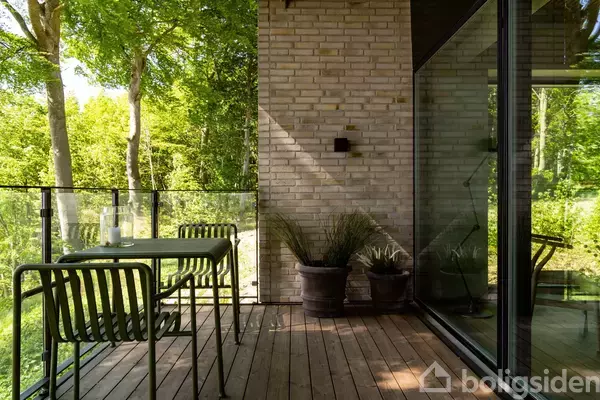 En terrasse med et bord og to stole står stille. Omkranset af glasvægge og grøn skovudsigter giver en fredfyldt atmosfære. Murstensvæggen tilføjer et moderne touch.