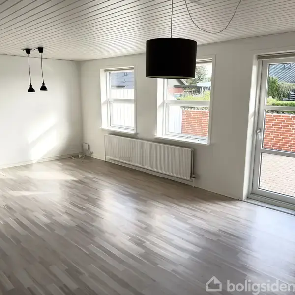 Tom stue med lyse trægulve og hvidt loft. To vinduer og en glasdør giver naturligt lys. Mørke loftslamper hænger i midten, med udsigt til en murstensvæg udenfor.