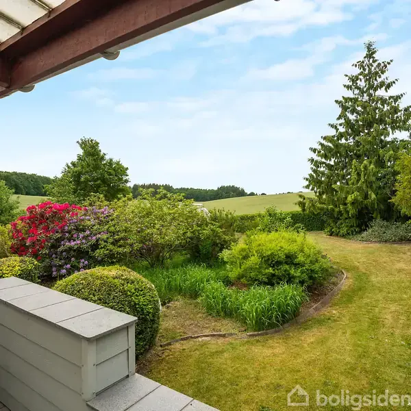 Terrasse under tagudhæng fører til frodig have med buske og blomsterbede. I baggrunden ses grønne marker og en klar, blå himmel.