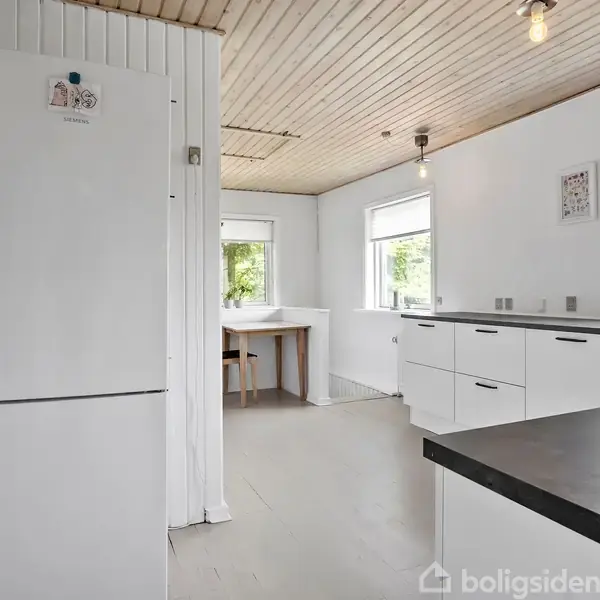 Køleskab står stille i et moderne, lyst køkken med hvide skabe. Træloft og vinduer giver naturligt lys, mens et bord er i baggrunden ved et vindue.