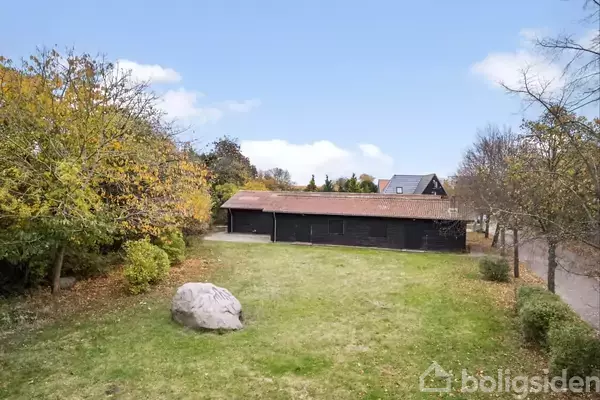 En stor sten ligger på en græsplæne foran en sort bygning med rødt tag. Omgivet af træer i et bebygget område.