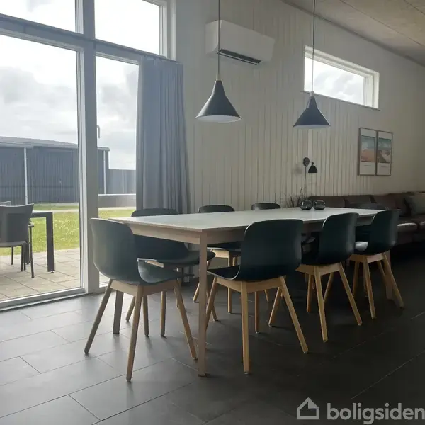 Et spisebord med sorte stole står i et lyst rum. En terrasse ses udenfor gennem store vinduer. To lamper hænger over bordet, og en sofa er i baggrunden.