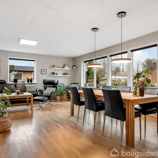 Stuen indeholder et spisebord med sorte stole og to loftslamper. Store vinduer giver udsigt til en have. En sofa og lænestole er placeret ved et sofabord med planter omkring.