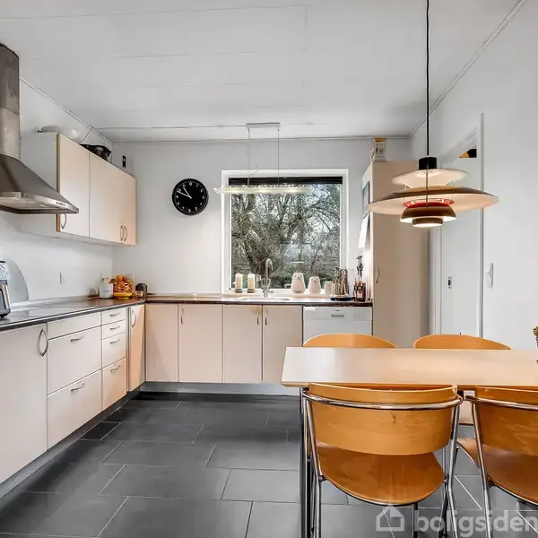 Køkkenbord med fire stole står i et lyst, moderne køkken med trægulve. En række skabe og skuffer danner køkkenets indretning. Et vindue fylder bagvæggen, og en vægur hænger ved siden af.