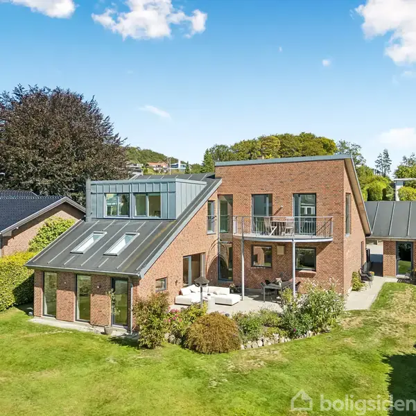En moderne murstensvilla med store vinduer står i et frodigt haveområde. Et balkonområde med havemøbler ses foran et grønt græsareal, omgivet af buske og træer.