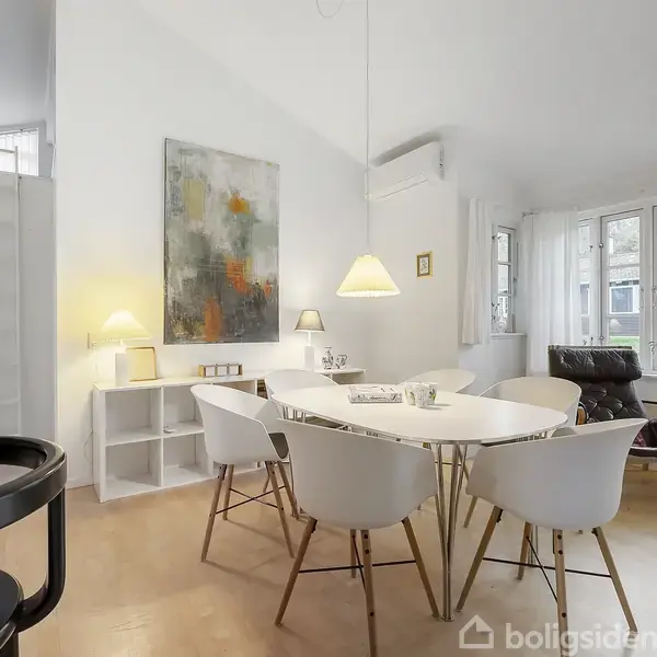 Fem hvide stole står rundt om et rundt bord i en lys stue med store vinduer. Over bordet hænger en lampe, og en farverig maleri pryder væggen.