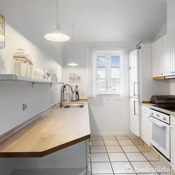 Et lyst køkken med lyse træbordplader, hvidt skabsarrangement og indbygget ovn. Der er en vask og vinduer for enden, som skaber et luftigt rum. En hylde på væggen.