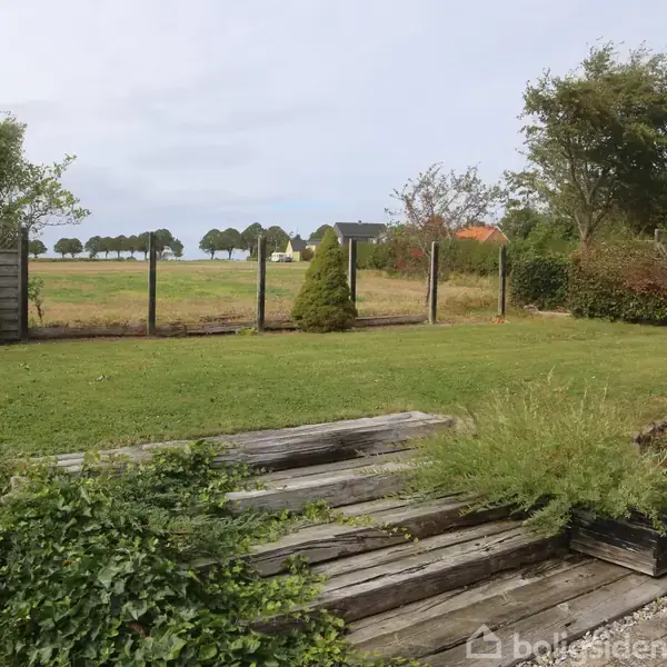 Sofa med puder står i en lys stue ved siden af et brunt bord med stor vase. Vinduet bagved har planter og billeder på hylden.