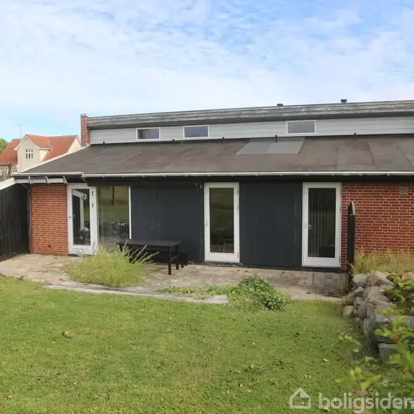 Et lille køkken med hvide skabe har en kaffemaskine og skåle på bordet. En trædør er placeret for enden af rummet, med køkkenredskaber til højre.