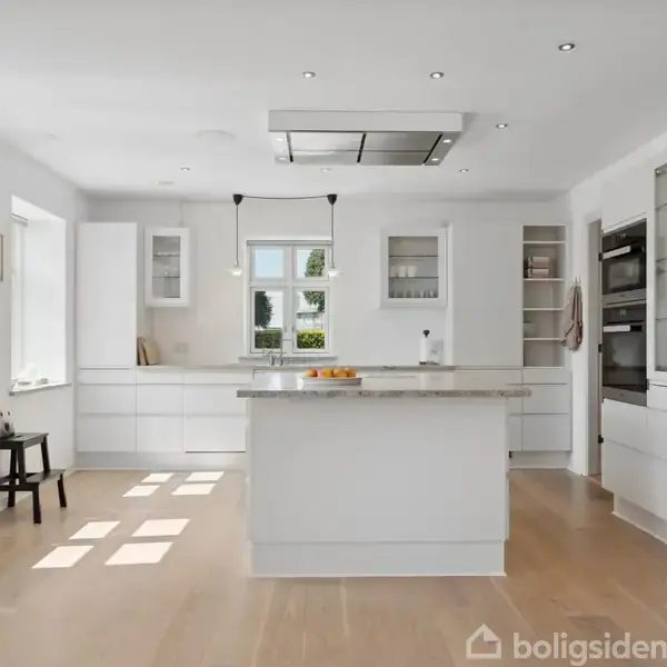 Et moderne køkken med hvide skabe og en stor ø i midten. Rummet er lyst med trægulv, store vinduer og en ovn indbygget i væggen. En stol står ved vinduet.