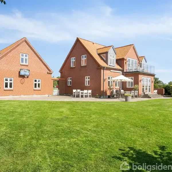 Et rødt murstenshus med tagterrasse står på en velplejet græsplæne. Huset har flere vinduer og udendørs møbler. Træer omkranser den solrige scene under en blå himmel.