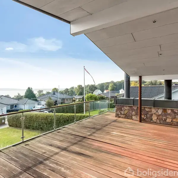 Stor terrasse med trægulv og glasrækværk, der står stille og skuer ud over et nabolag med huse samt en fjern udsigt til vand og skovklædte områder under en blå himmel.