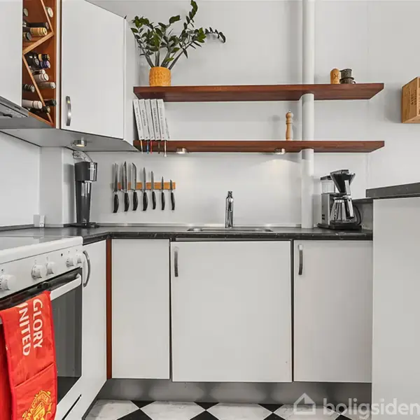 Et hvidt køkken med sort bordplade. Ovnen har et rødt håndklæde med teksten "GLORY UNITED". Knive hænger på væggen. Over vasken er der træhylder med planter og bøger.